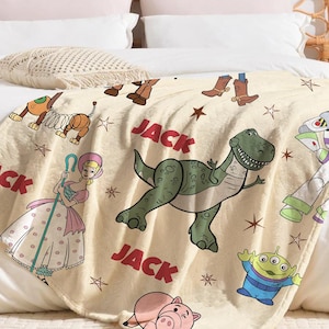 May include: A beige throw blanket featuring cartoon characters from the movie Toy Story. The blanket has images of Rex the dinosaur, Buzz Lightyear, and other characters. The word "JACK" is printed in red on the blanket.