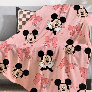 Op de afbeelding: Roze plaid met een herhalend patroon van Mickey Mouse, roze strikken, bloemen en rode sterren. De deken is over een grijze bank gedrapeerd en toont de zachte, pluche textuur en het levendige ontwerp.
