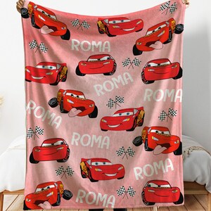 Puede incluir: Una manta rosa y roja con un patr&oacute;n repetido de un coche de carreras de dibujos animados rojo, banderas a cuadros y la palabra "ROMA". La manta est&aacute; sujeta por una persona.