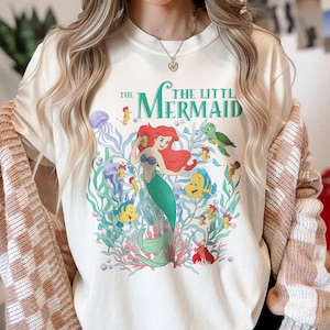 Puede incluir: Camiseta de color marfil con un gr&aacute;fico colorido de Ariel de La Sirenita. El dise&ntilde;o incluye a Ariel con cabello rojo y cola verde, rodeada de criaturas marinas y el texto "The Little Mermaid".