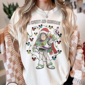 May include: Ivory-coloured t-shirt with a graphic of Buzz Lightyear in a Santa hat, encircled by Mickey Mouse-shaped Christmas lights. The text "TO INFINITY AND CHRISTMAS!" is printed above the graphic. This festive shirt is part of a holiday collection.