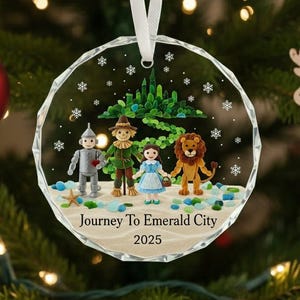 May include: A clear, round Christmas ornament with a detailed scene from The Wizard of Oz. The ornament features the Tin Man, Scarecrow, Dorothy, and the Cowardly Lion in front of the Emerald City. The text "Journey To Emerald City 2025" is printed on the bottom.