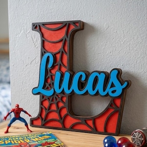 May include: A large wooden letter L featuring a red background with a black spiderweb overlay and the name Lucas in blue script lettering. The item is displayed alongside a Spider-Man action figure, a comic book, and small spheres.