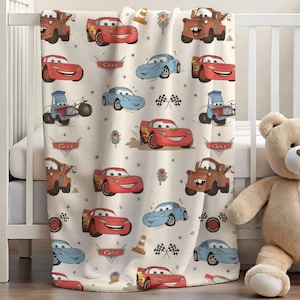 Puede incluir: Una manta de beb&eacute; color crema con un patr&oacute;n repetido de coches animados en rojo, azul y marr&oacute;n. La manta presenta im&aacute;genes de banderas de carreras, sem&aacute;foros y la palabra "Cars". Un oso de peluche est&aacute; al lado de la cuna.