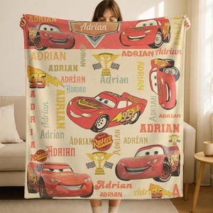 Op de afbeelding: Een beige deken met Lightning McQueen uit de film Cars. De deken heeft een herhalend patroon van het personage, het nummer 95 en de naam "Adrian" in rood en geel. Geschikt voor bed of bank.