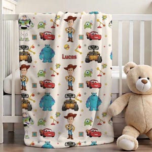 May include: A cream-colored baby blanket featuring cartoon characters from the movie Toy Story and Monsters, Inc. The blanket has images of Woody, Buzz Lightyear, Sully, and others. The name "Lucas" is printed on the blanket.