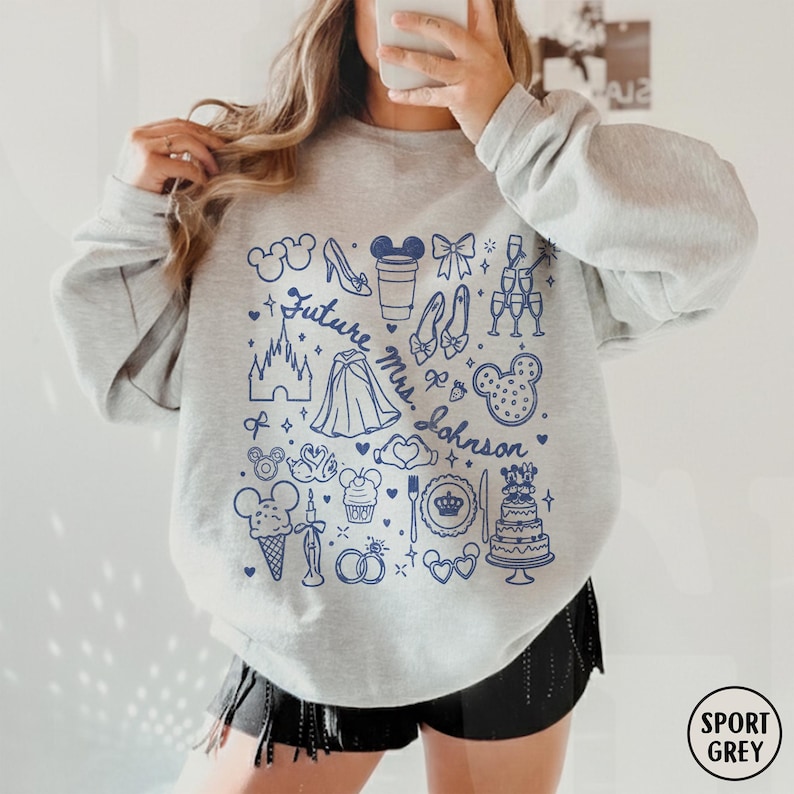 May include: A light grey crewneck sweatshirt featuring a dark blue graphic print of various wedding and fairy tale icons, including a castle, rings, a cake, and the text Future Mrs. Johnson, arranged in a collage pattern across the front chest.