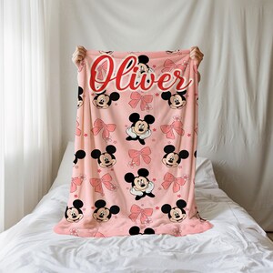 Op de afbeelding: Roze deken met een patroon van Mickey Mouse-gezichten, roze strikken en bloemen. De naam "Oliver" staat in sierlijke letters bovenaan. De deken wordt gepresenteerd tegen een witte achtergrond.