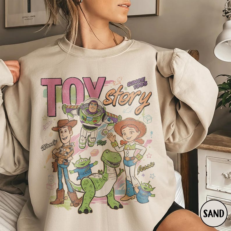 May include: Beige sweatshirt featuring a colorful graphic of characters from the animated film "Toy Story." The design includes Woody, Buzz Lightyear, Jessie, and other characters, with the words "Toy Story" and "Since 1995."