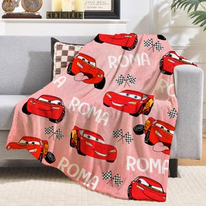 Puede incluir: Una manta rosa con un estampado de coches de carreras rojos de dibujos animados, banderas a cuadros y la palabra "ROMA" en blanco. La manta est&aacute; sobre un sof&aacute; gris. Los coches tienen caras sonrientes y est&aacute;n en varias poses.