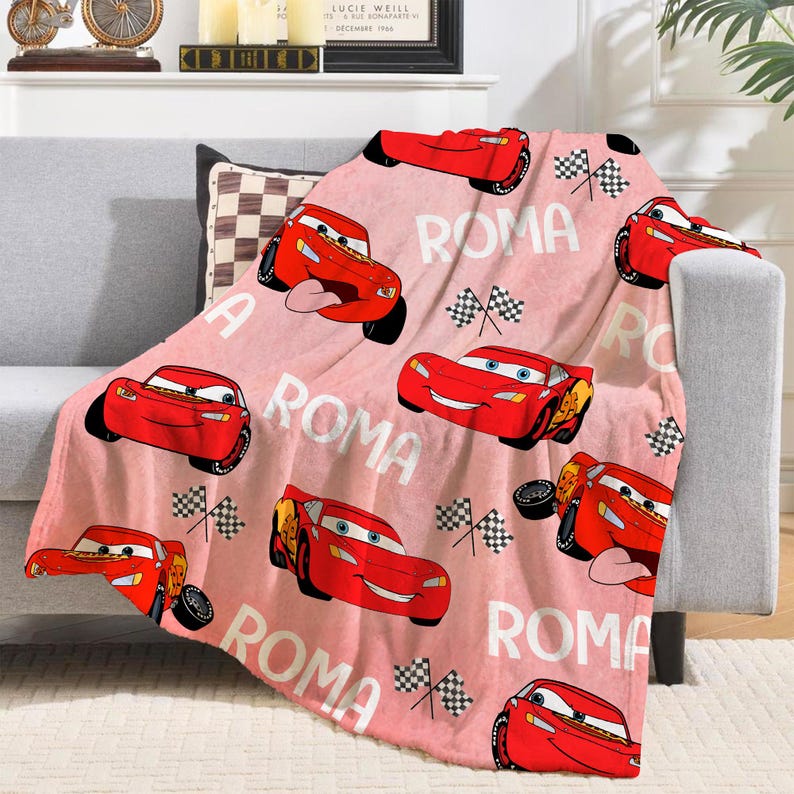 Puede incluir: Una manta rosa con un estampado de coches de carreras rojos de dibujos animados, banderas a cuadros y la palabra "ROMA" en blanco. La manta est&aacute; sobre un sof&aacute; gris. Los coches tienen caras sonrientes y est&aacute;n en varias poses.
