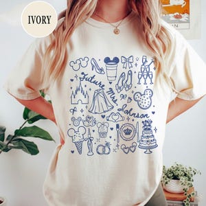 May include: An ivory t-shirt featuring a blue graphic print with the text "Future Mrs. Johnson" surrounded by various icons including a castle, a wedding cake, champagne glasses, mouse ears, a glass slipper, and other wedding-themed illustrations.