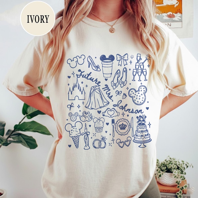 May include: An ivory t-shirt featuring a blue graphic print with the text "Future Mrs. Johnson" surrounded by various icons including a castle, a wedding cake, champagne glasses, mouse ears, a glass slipper, and other wedding-themed illustrations.
