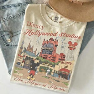Può includere: T-shirt color crema con grafica Disney Hollywood Studios. Il design include Topolino, la Torre del Terrore e il testo "The Magic of Movies". La maglietta è piegata sopra jeans blu, con una collana e un cappello visibili.