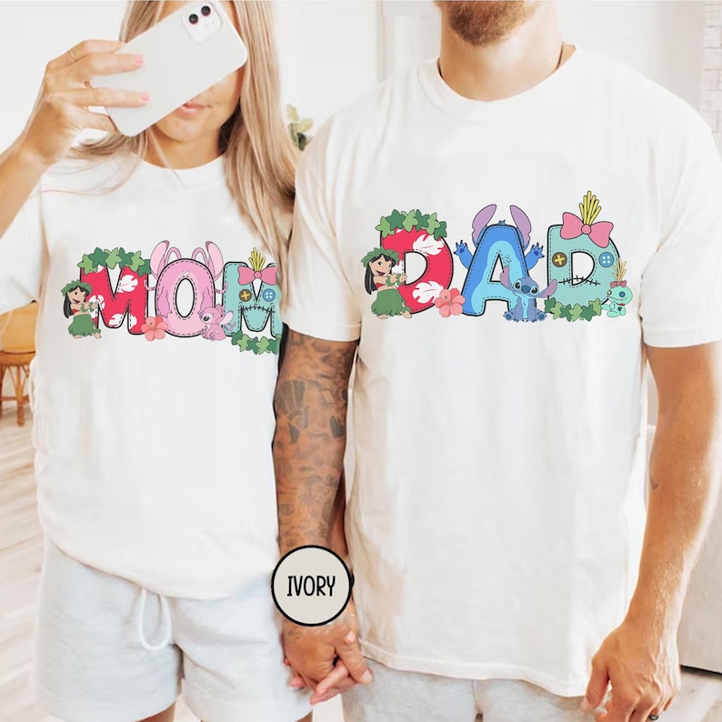 K&ouml;nnte beinhalten: Elfenbeinfarbene T-Shirts mit farbenfrohen Grafiken. Eines zeigt "MOM" mit floralen und Charakter-Akzenten. Das andere zeigt "DAD" mit &auml;hnlichen Designelementen. Die Shirts sind wahrscheinlich f&uuml;r eine Familie.