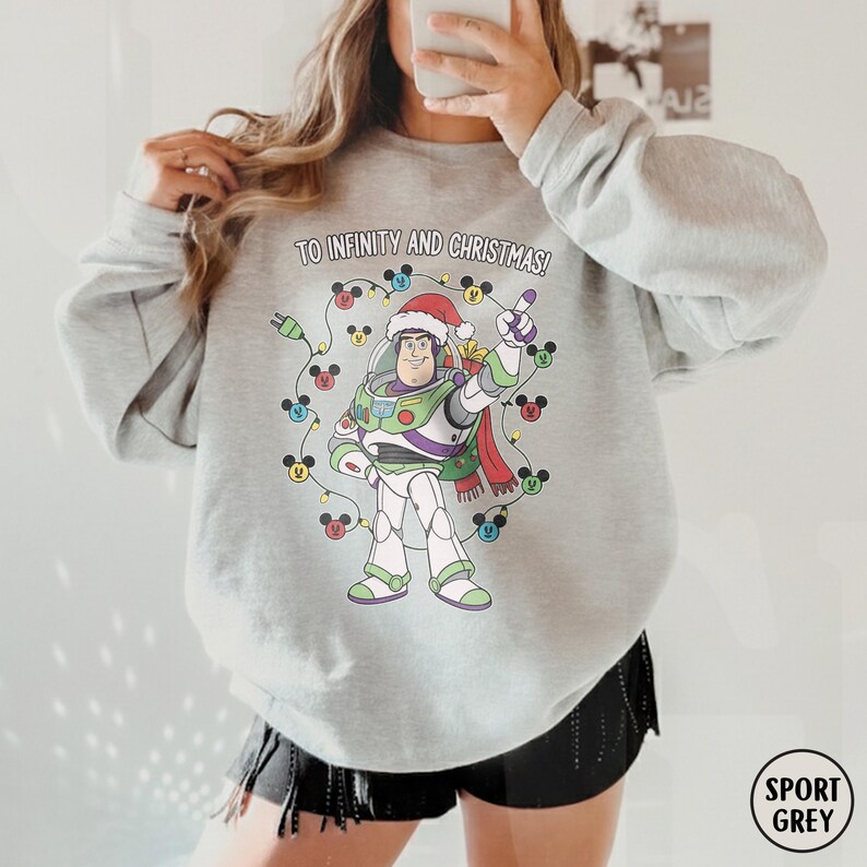May include: A light grey sweatshirt with a Buzz Lightyear graphic wearing a Santa hat, encircled by Mickey Mouse Christmas lights. The text "TO INFINITY AND CHRISTMAS!" is printed above the character. The sweatshirt is labelled "SPORT GREY".