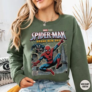 K&ouml;nnte beinhalten: Milit&auml;rgr&uuml;nes Sweatshirt mit einem Comic-Stil-Grafik von Spider-Man und Venom. Der Text auf dem Sweatshirt lautet "SPIDER-MAN BRAND NEW DAY" und "DAILY BUGLE". Das Sweatshirt hat auch ein Spider-Man-Logo.