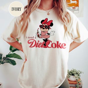 May include: Ivory-colored t-shirt featuring a cartoon character wearing heart-shaped sunglasses and a pink dress, holding a Diet Coke bottle. The text "i need a Diet Coke" is printed in red script.