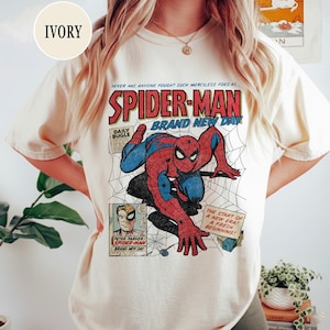 Puede incluir: Camiseta de color marfil con un diseño de cómic vintage de Spider-Man. El gráfico muestra a Spider-Man en acción, con detalles de telaraña y el texto "SPIDER-MAN BRAND NEW DAY". La camiseta tiene un corte clásico y es cómoda.