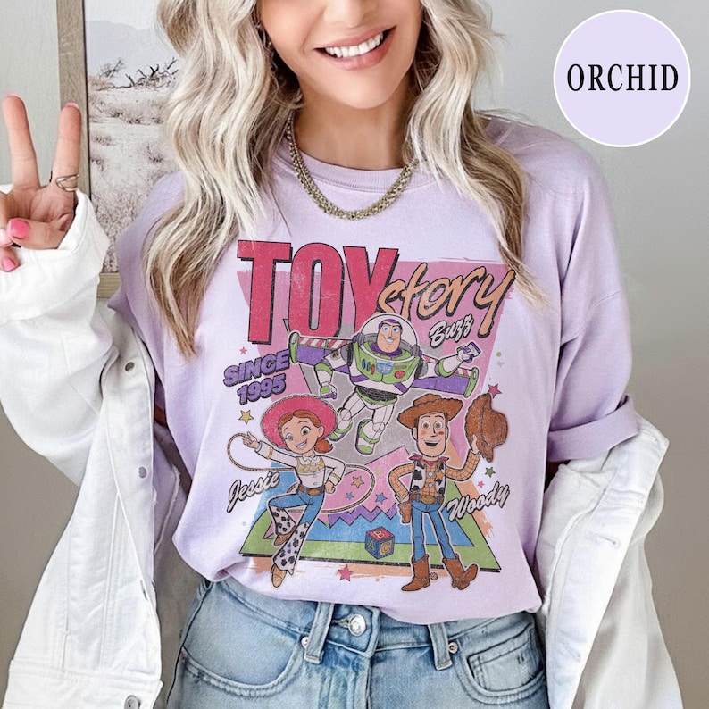 May include: A lilac long-sleeved shirt with a Toy Story graphic featuring Buzz Lightyear, Jessie, and Woody. The shirt displays the text "Toy Story" and "Since 1995". The shirt is a relaxed, casual style.