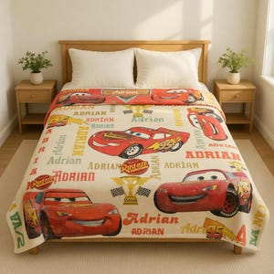 Op de afbeelding: Een beige deken met het personage Lightning McQueen uit de film Cars. De deken heeft een herhalend patroon van het personage, het woord "Adrian" en graphics met racethema. De deken ligt op een houten bedframe.