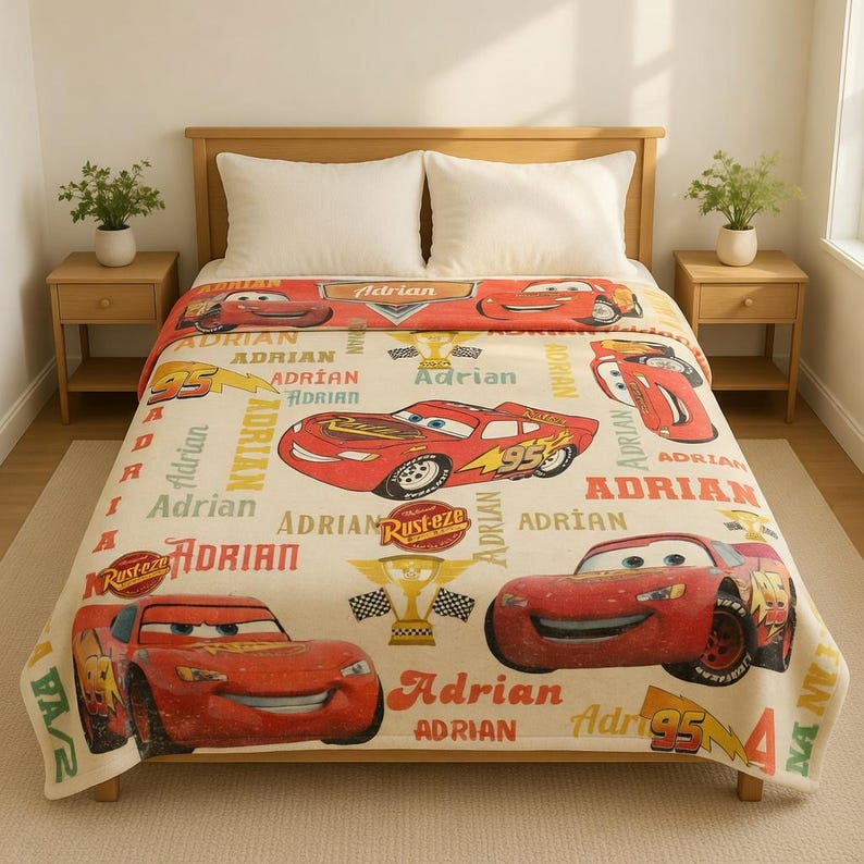 Op de afbeelding: Een beige deken met het personage Lightning McQueen uit de film Cars. De deken heeft een herhalend patroon van het personage, het woord "Adrian" en graphics met racethema. De deken ligt op een houten bedframe.