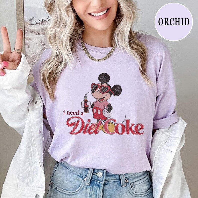 May include: A light purple t-shirt featuring a graphic of Mickey Mouse wearing sunglasses and holding a Diet Coke. The text "I need a Diet Coke" is printed below the cartoon character in a retro font.