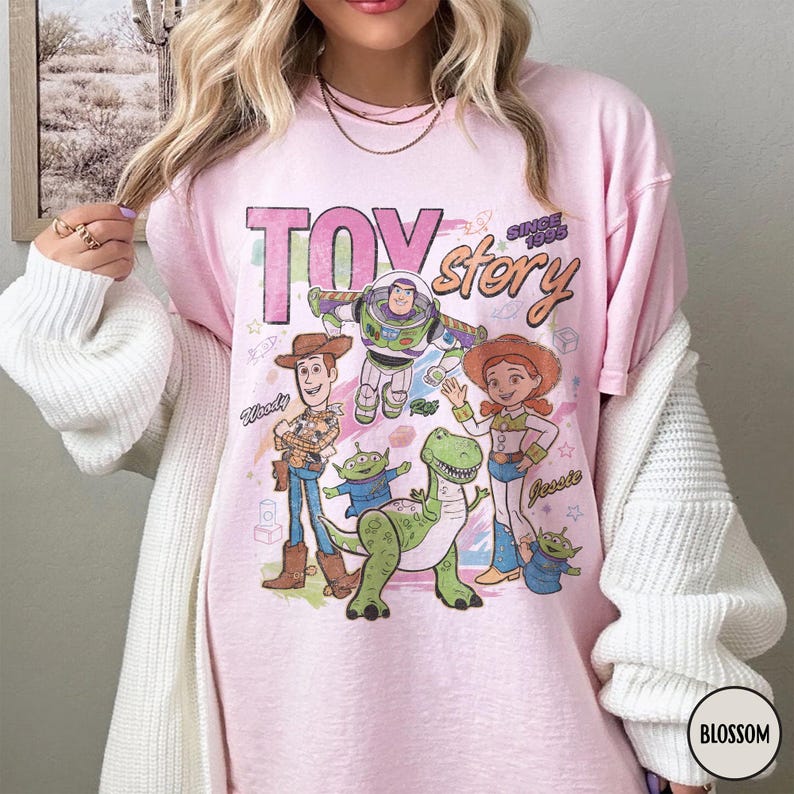 May include: Pink t-shirt featuring the words "Toy Story" with the phrase "Since 1995" and images of Woody, Buzz Lightyear, Jessie, a green dinosaur, and two green aliens. The shirt has a vintage, distressed look.