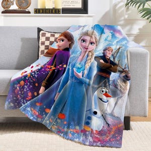 K&ouml;nnte beinhalten: Eine weiche Kuscheldecke mit Figuren aus dem Animationsfilm "Die Eisk&ouml;nigin". Die Decke zeigt Elsa, Anna, Kristoff, Olaf und Sven vor einer schneebedeckten Kulisse. Die Decke ist &uuml;berwiegend blau, lila und wei&szlig;.