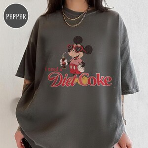 May include: Dark gray t-shirt featuring a vintage-style graphic of Mickey Mouse wearing heart-shaped sunglasses and holding a Diet Coke. The text "I need a Diet Coke" is printed below the character in a retro font.