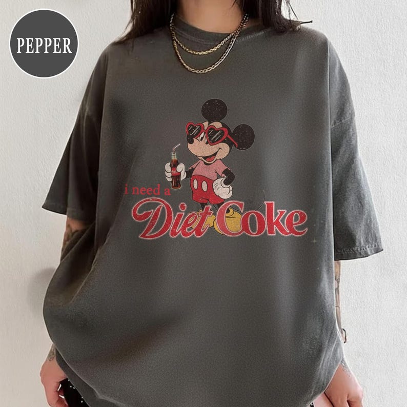 May include: Dark gray t-shirt featuring a vintage-style graphic of Mickey Mouse wearing heart-shaped sunglasses and holding a Diet Coke. The text "I need a Diet Coke" is printed below the character in a retro font.