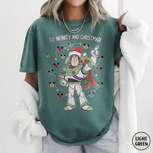 May include: Light green t-shirt with a graphic of Buzz Lightyear in a Santa hat, surrounded by Mickey Mouse lights. The text "TO INFINITY AND CHRISTMAS!" is printed above the character. The shirt has a relaxed fit.
