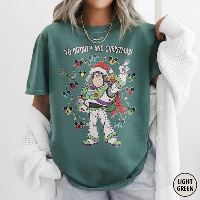May include: Light green t-shirt with a graphic of Buzz Lightyear in a Santa hat, surrounded by Mickey Mouse lights. The text "TO INFINITY AND CHRISTMAS!" is printed above the character. The shirt has a relaxed fit.