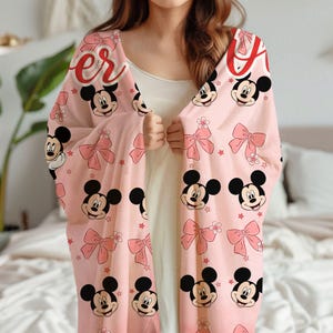 Op de afbeelding: Een lichtroze deken met Mickey Mouse gezichten, roze strikken en rode sterren. Het woord "Forever" is in rode letters gedrukt. Een decoratief item voor thuis.