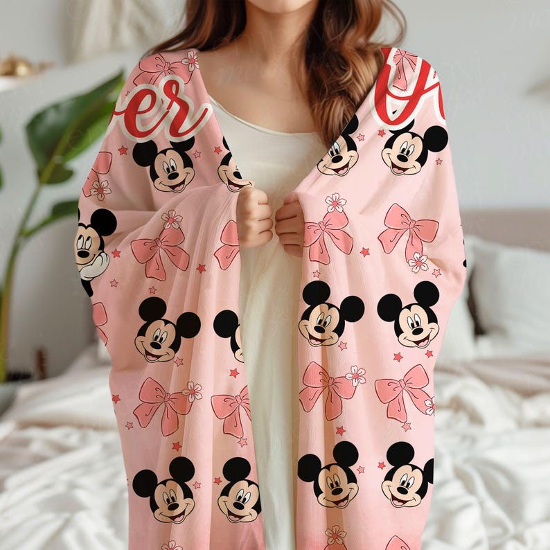 Op de afbeelding: Een lichtroze deken met Mickey Mouse gezichten, roze strikken en rode sterren. Het woord "Forever" is in rode letters gedrukt. Een decoratief item voor thuis.