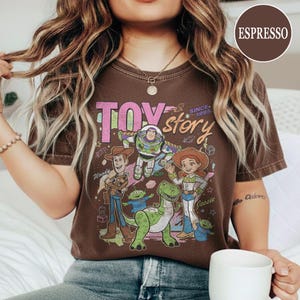 May include: Espresso-colored t-shirt featuring a graphic of Toy Story characters, including Woody, Buzz Lightyear, and Jessie. The words "TOY STORY" and "SINCE 1995" are printed on the front. The shirt is a crew neck.