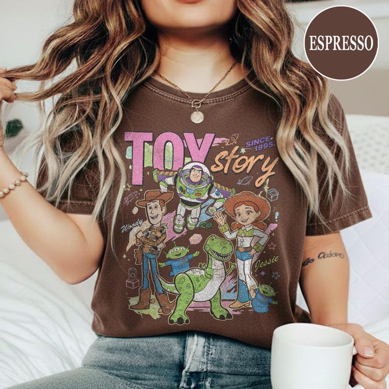May include: Espresso-colored t-shirt featuring a graphic of Toy Story characters, including Woody, Buzz Lightyear, and Jessie. The words "TOY STORY" and "SINCE 1995" are printed on the front. The shirt is a crew neck.