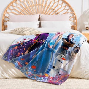 K&ouml;nnte beinhalten: Eine weiche Decke mit Figuren aus dem Animationsfilm Frozen. Die Decke zeigt Elsa in einem blauen Kleid, Anna in einem lila Kleid, Kristoff, Olaf den Schneemann und Sven das Rentier. Die Decke liegt auf einem wei&szlig;en Bett.