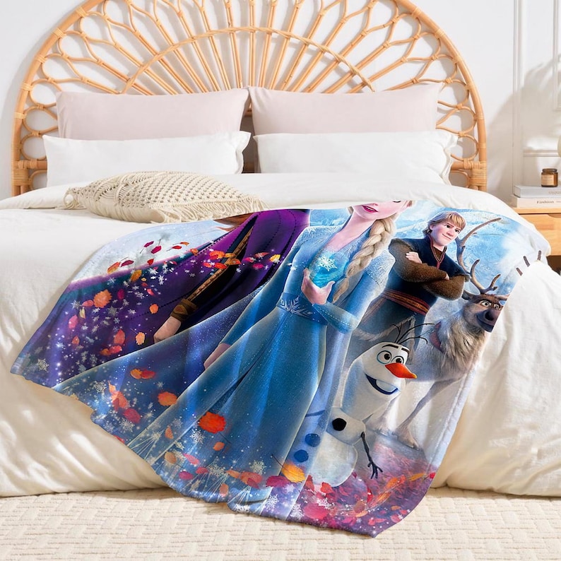 K&ouml;nnte beinhalten: Eine weiche Decke mit Figuren aus dem Animationsfilm Frozen. Die Decke zeigt Elsa in einem blauen Kleid, Anna in einem lila Kleid, Kristoff, Olaf den Schneemann und Sven das Rentier. Die Decke liegt auf einem wei&szlig;en Bett.