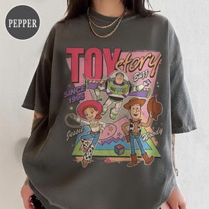 May include: Dark grey t-shirt with a Toy Story graphic featuring Buzz Lightyear, Woody, and Jessie. The text "TOY STORY" is in pink, with "Buzz" in white. The shirt also includes the text "Since 1995".