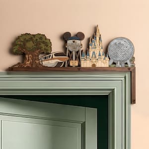 Peut inclure: Un ornement de porte en bois présentant un design superposé avec un arbre, un monorail, un château d'eau avec le texte WDI 1935, un château et un dôme géodésique. L'étagère en bois foncé comporte des découpes en forme de tête de Mickey.