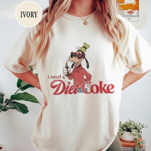K&ouml;nnte beinhalten: Elfenbeinfarbenes T-Shirt mit einem Retro-Grafikdesign eines Zeichentrickhundes, der eine Sonnenbrille tr&auml;gt und aus einer Diet Coke-Flasche trinkt. Der Text "i need a Diet Coke" ist unten in einer Vintage-Schriftart aufgedruckt.