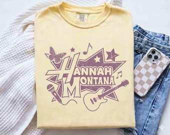 May include: A pale yellow t-shirt with a purple graphic featuring the words "Hannah Montana". The design includes a butterfly, musical notes, a star, a microphone, and a guitar. The shirt is folded on a white blanket.