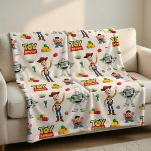 May include: A cream-colored throw blanket featuring a repeating pattern of characters and elements from the animated film "Toy Story." The design includes Woody, Buzz Lightyear, Mr. Potato Head, and the text "Toy Story."