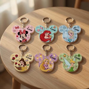 May include: Six Mickey-shaped keychains featuring Disney characters: Minnie Mouse, Ariel, Elsa, Belle, Rapunzel, and Tinker Bell. Each keychain has a metal ring and is personalized with the name Luna or Euna printed in black script on the surface.