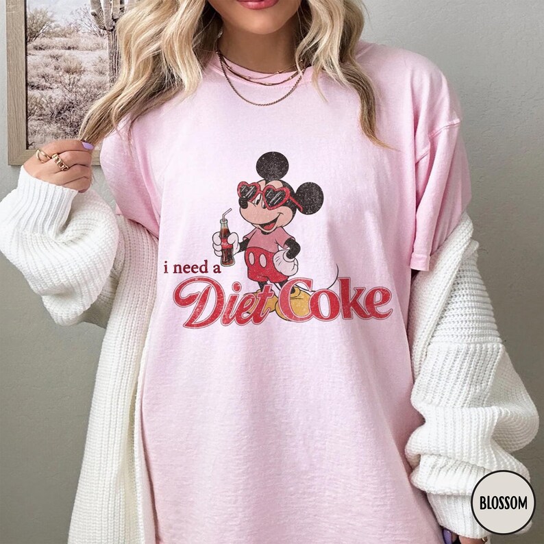 May include: Light pink t-shirt featuring a graphic of Mickey Mouse wearing heart-shaped sunglasses and holding a Diet Coke. The text "i need a Diet Coke" is printed below the graphic in a retro font.