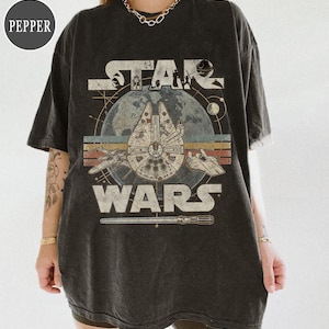 Könnte beinhalten: Dunkelgraues T-Shirt mit einem Star Wars-Motiv. Das Design zeigt den Text "STAR WARS" mit Bildern des Millennium Falcon, Raumschiffen und einem Lichtschwert. Die Grafik ist vor einem blauen Planetenhintergrund mit Retro-Streifen platziert.
