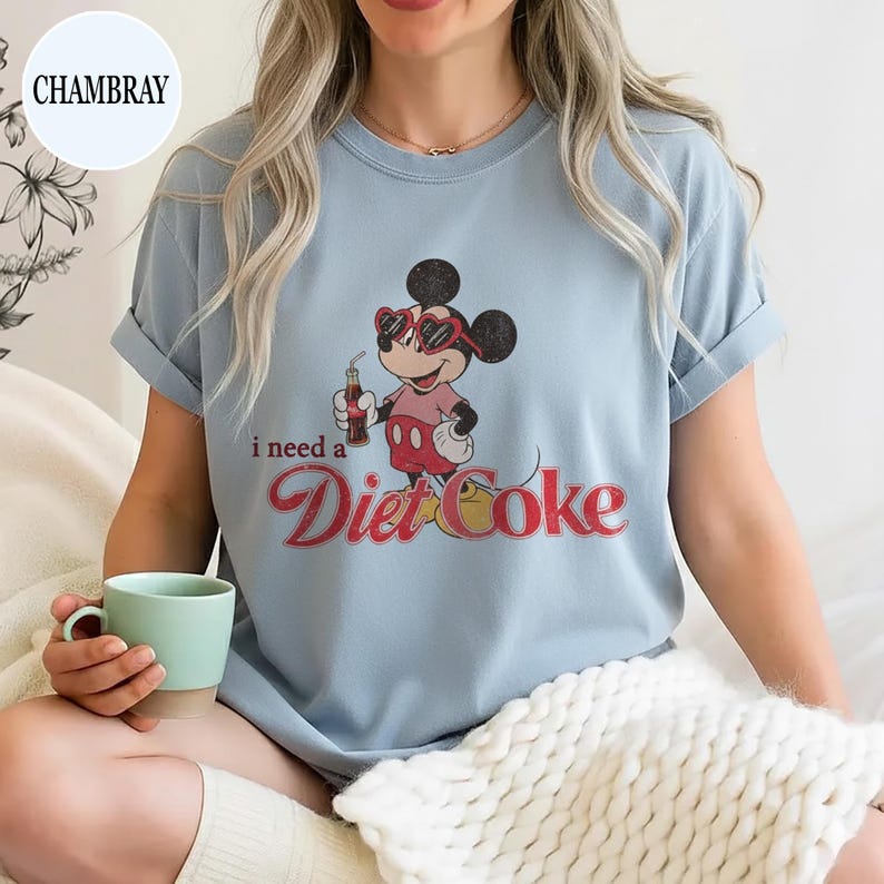 May include: Light blue t-shirt featuring a vintage-style graphic of Mickey Mouse wearing sunglasses and holding a Diet Coke. The text "I need a Diet Coke" is printed below the graphic in a retro font. The shirt has rolled-up sleeves.
