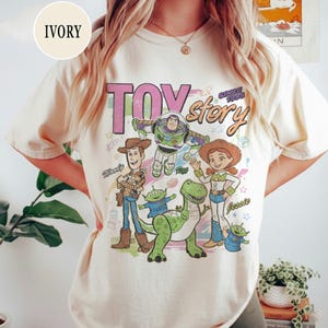 May include: Ivory-colored t-shirt featuring a graphic of Toy Story characters, including Woody, Buzz Lightyear, and Jessie. The text "TOY STORY" and "SINCE 1995" are printed above the characters. The shirt is a casual, crew neck style.