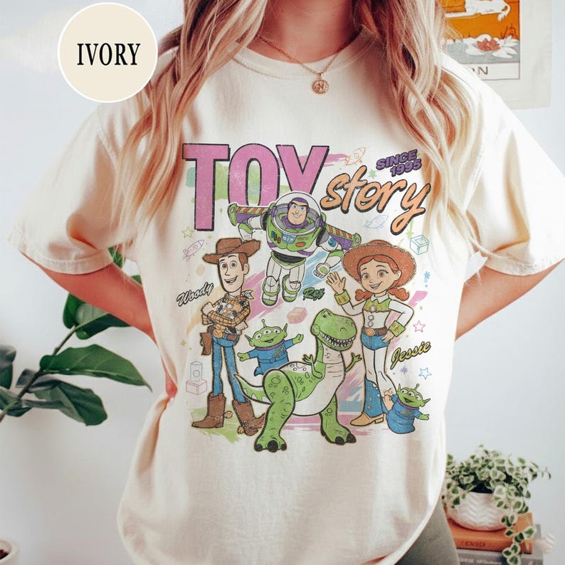 May include: Ivory-colored t-shirt featuring a graphic of Toy Story characters, including Woody, Buzz Lightyear, and Jessie. The text "TOY STORY" and "SINCE 1995" are printed above the characters. The shirt is a casual, crew neck style.